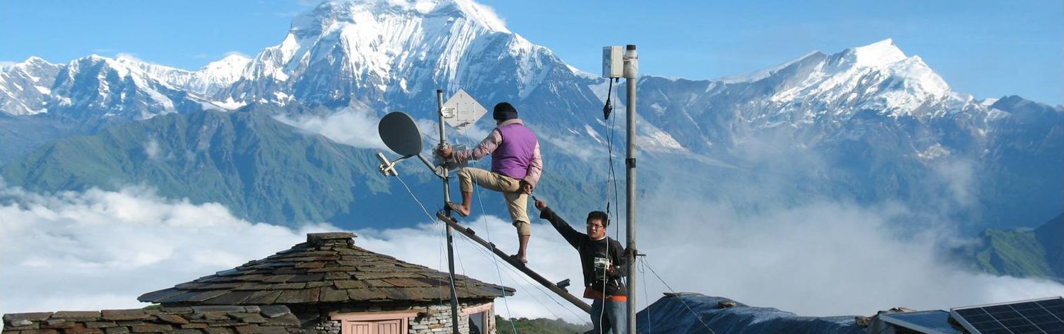 Two men fitting antennae on top of a room in front of the Himalaya mountains in Nepal