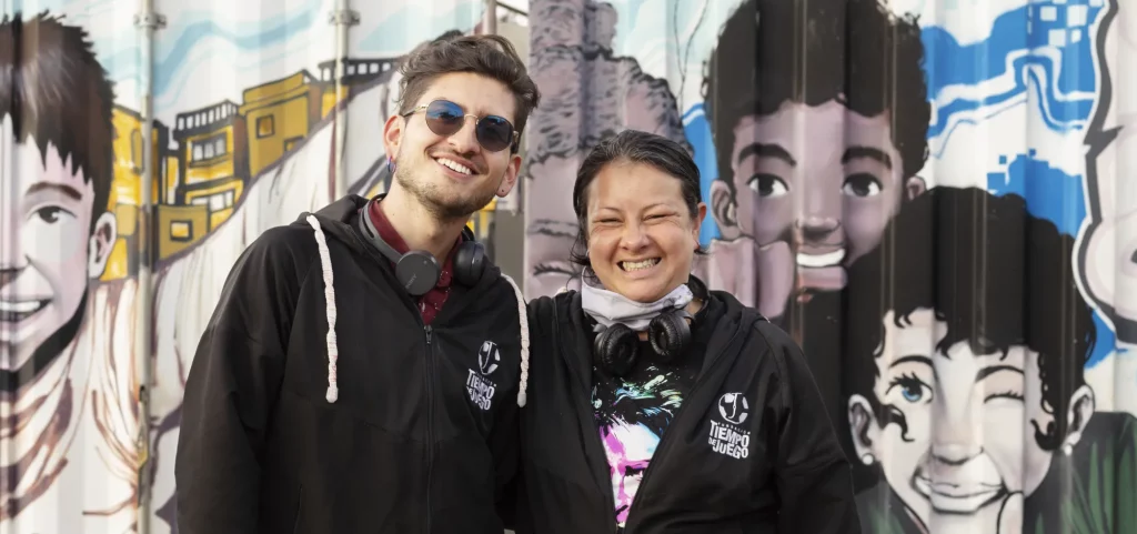 Two people from Tiempo del Juego stand in front of a graffiti covered wall