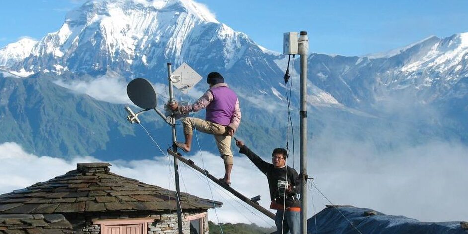 Two men fitting antennae on top of a room in front of the Himalaya mountains in Nepal