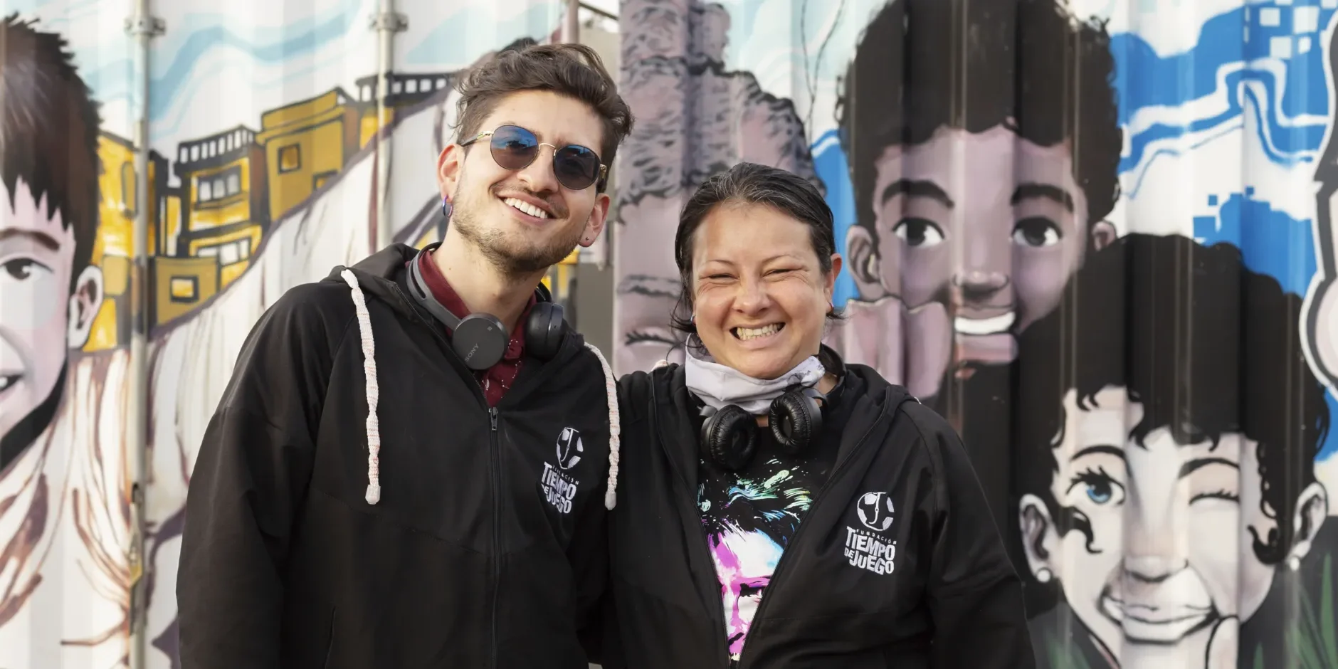 Two people from Tiempo del Juego stand in front of a graffiti covered wall